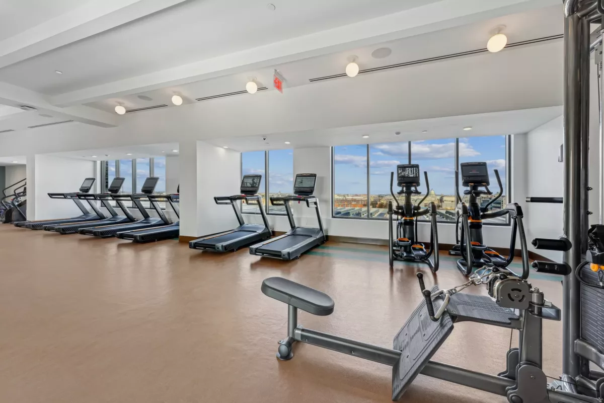 Fitness center with treadmills, ellipticals, white walls, and large windows with city views.
