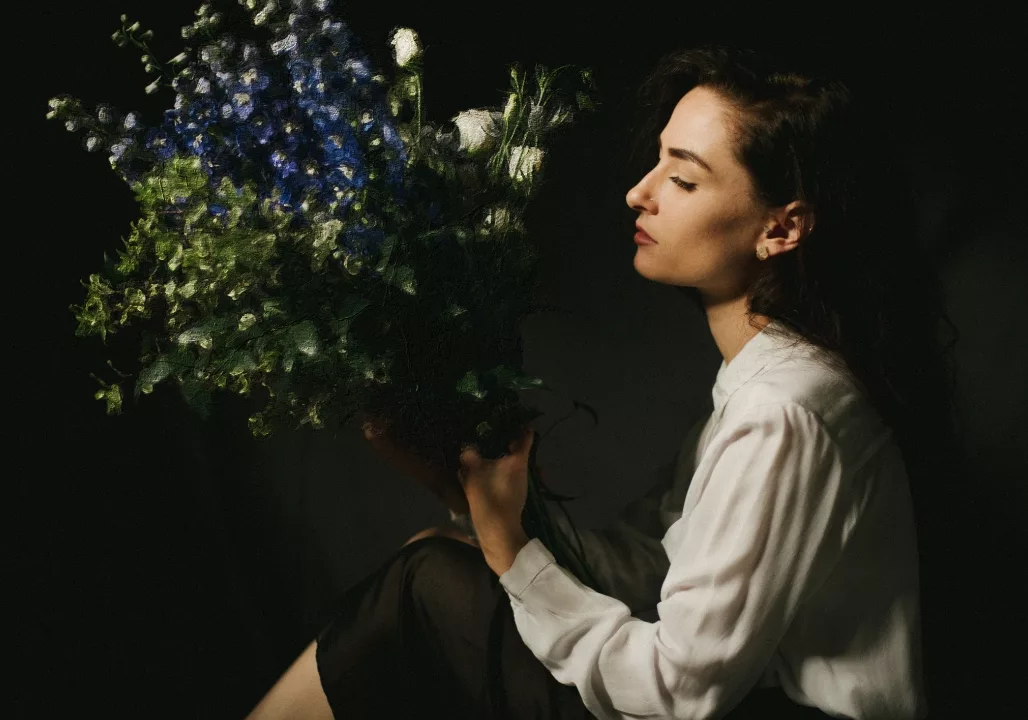 Woman in a white blouse holding a large bouquet of flowers, set against a dark background.