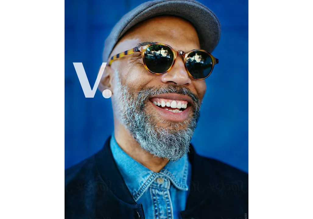 Smiling man with glasses, a gray beard, and a cap, wearing a blue shirt, against a blue background with a "V." on top.