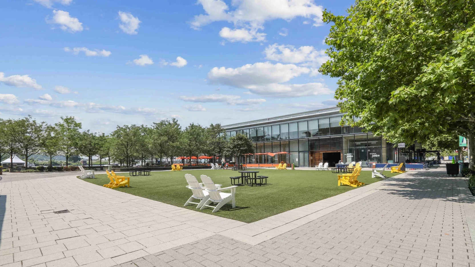 Outdoor plaza with green turf, surrounded by trees, and featuring colorful chairs and tables under a blue sky.