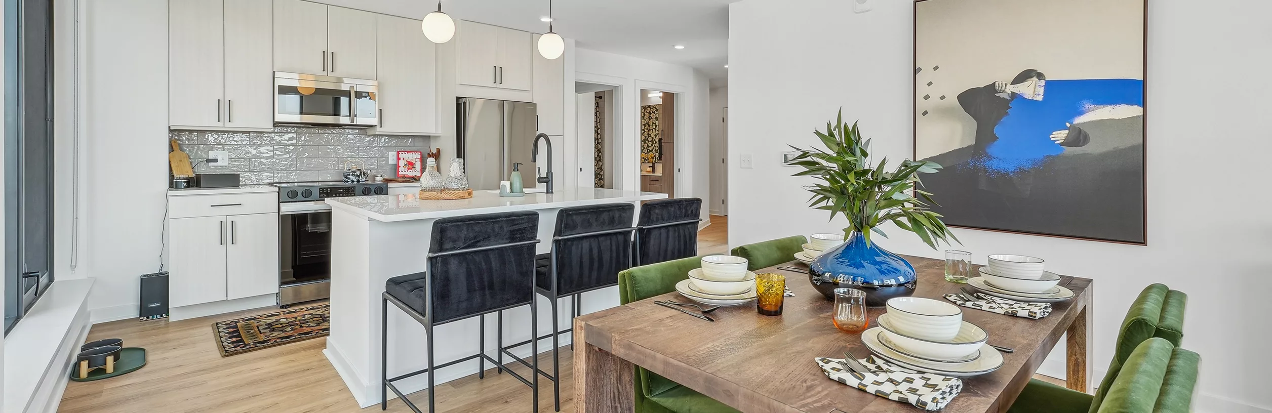 Open-concept kitchen and dining area with a kitchen island with black bar stools, a wooden table with green chairs, and modern wall art.