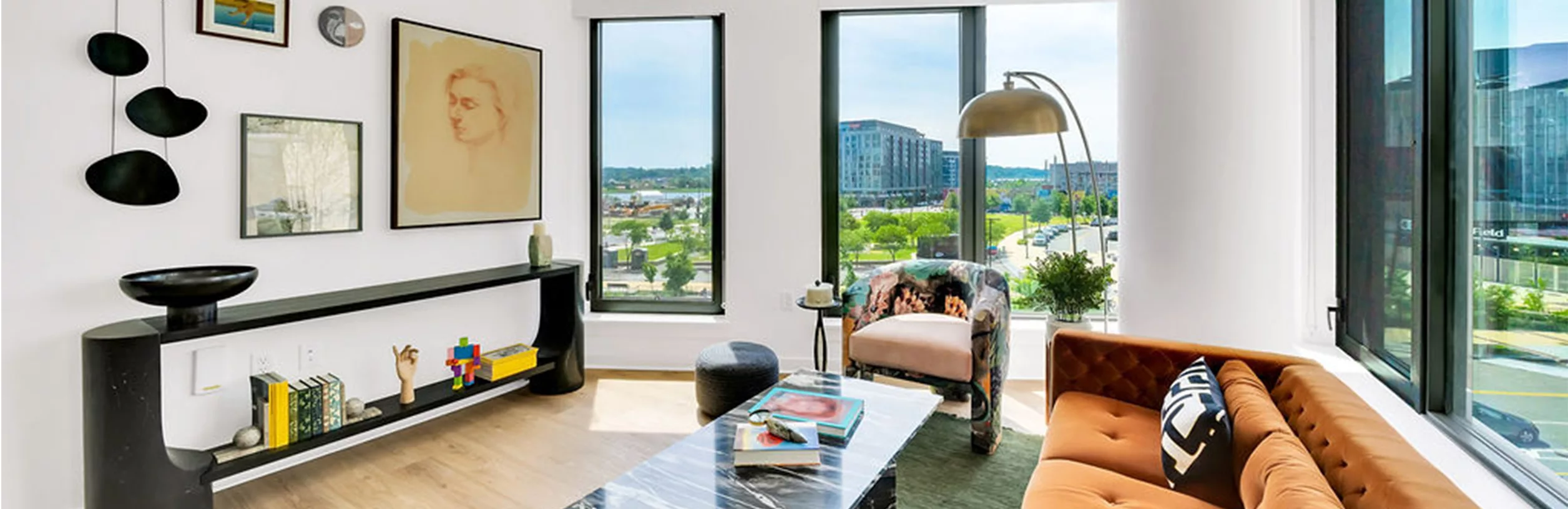 A living room with an orange sofa, patterned armchair, coffee table with books, modern art on the wall, and large windows.